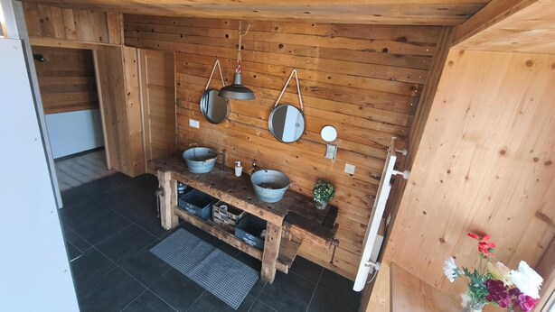 Un interior de madera con un banco de madera, dos cuencos metálicos, un espejo, una lámpara colgante y una pequeña planta.