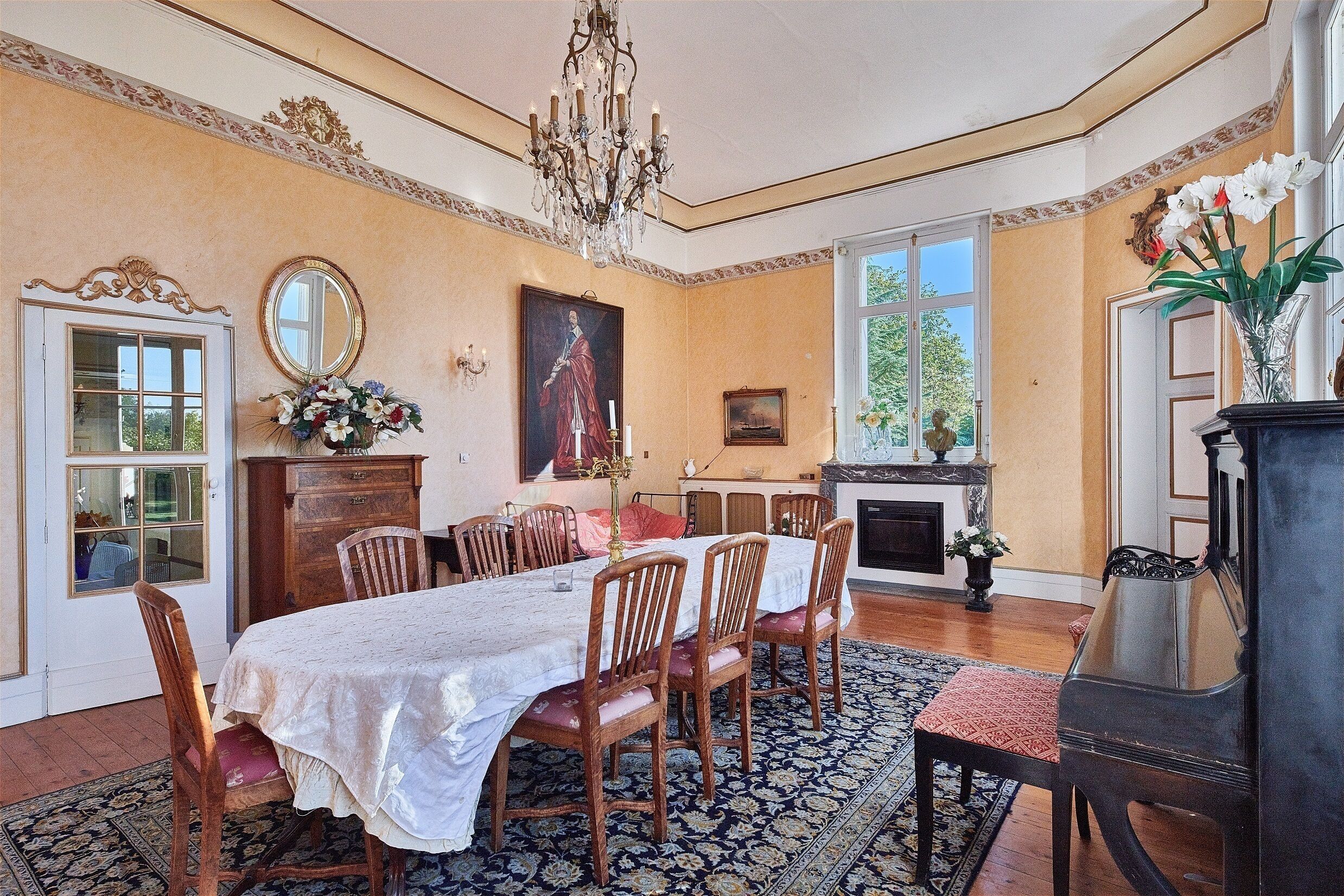 A dining room with a large table, chairs, a chandelier, a piano, and a fireplace.