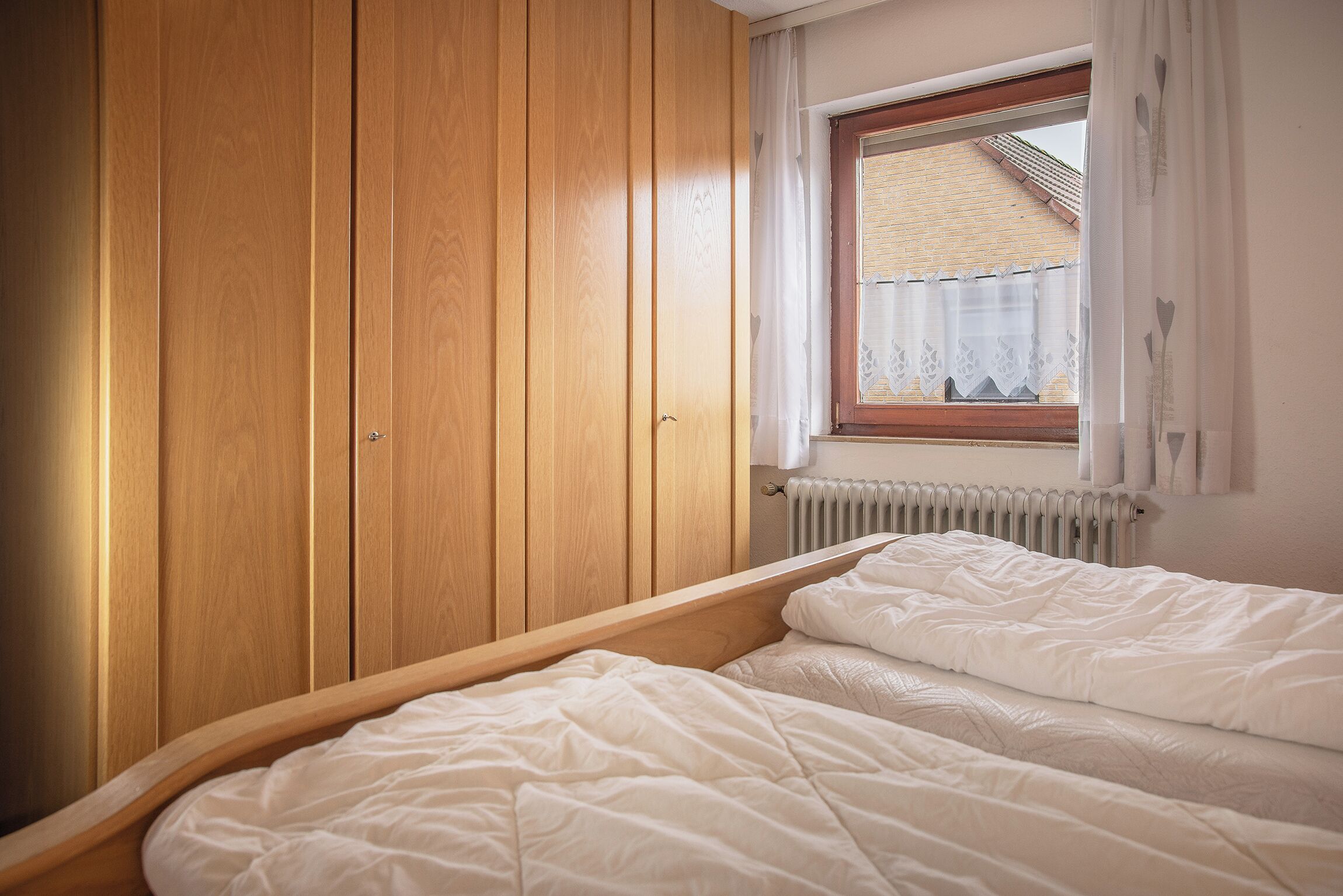 A bedroom with a wooden wardrobe, a window with a view of a brick building, and a radiator.