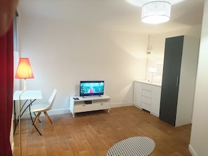 Ett modernt vardagsrum med en platt-TV, ett vitt TV-bord, ett litet vitt bord med en stol, en röd lampa och en mönstrad matta.
