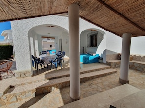A covered outdoor seating area with a table and chairs, a blue sofa, and a staircase leading to an upper level.