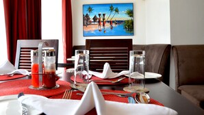 A dining table set with red placemats, clear glasses, and silverware, with a painting of a beach scene on the wall behind. - Villa Machakos Hotel and Conferencing (Machakos)