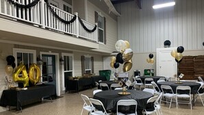 A spacious indoor venue with multiple round tables and white chairs, decorated with black and gold balloons, suggesting a celebration or event.