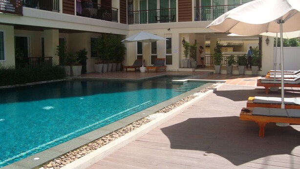 A pool area with lounge chairs and umbrellas, surrounded by potted plants and a building with balconies. - SUPER 404 LUXURY APARTMENT, QUIET, IN THE DINAMIQUE DISTRICT OF JOMTIEN. (Jomtien)