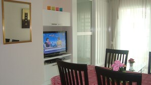 A living room with a TV, a dining table with chairs, and a window with curtains. - SUPER 404 LUXURY APARTMENT, QUIET, IN THE DINAMIQUE DISTRICT OF JOMTIEN. (Jomtien)