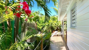 Une terrasse en bois menant à une maison dotée d’un porche abrité, le tout entouré d’une végétation tropicale luxuriante.