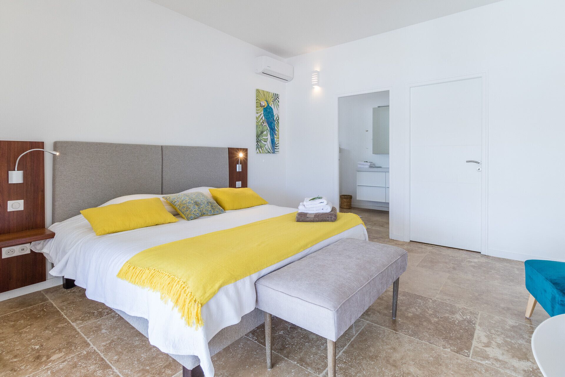A hotel room with a bed, bedside table, bench, and a bathroom visible through an open door.