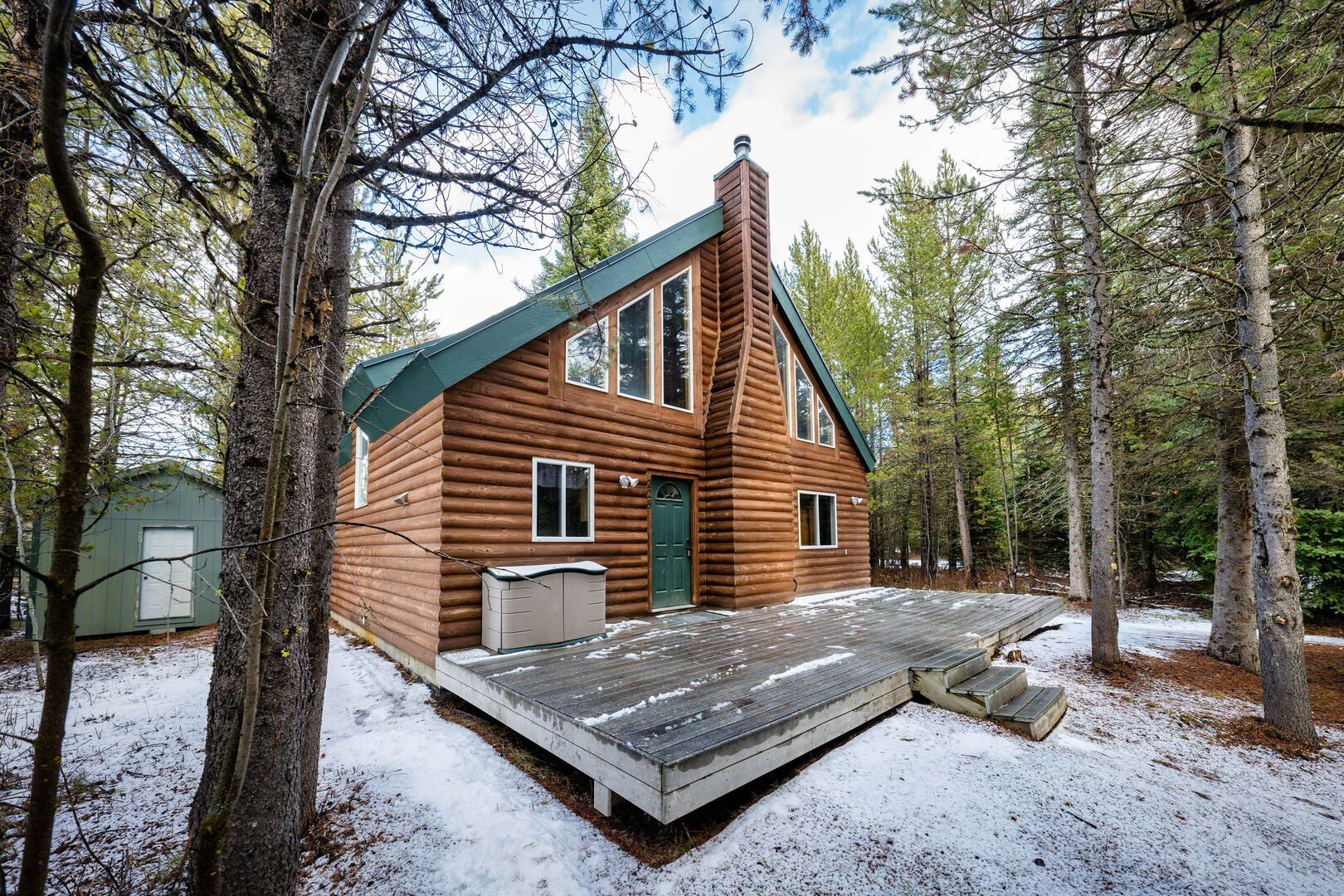 A log cabin with a green roof, a front porch, and a small shed in a forested area.