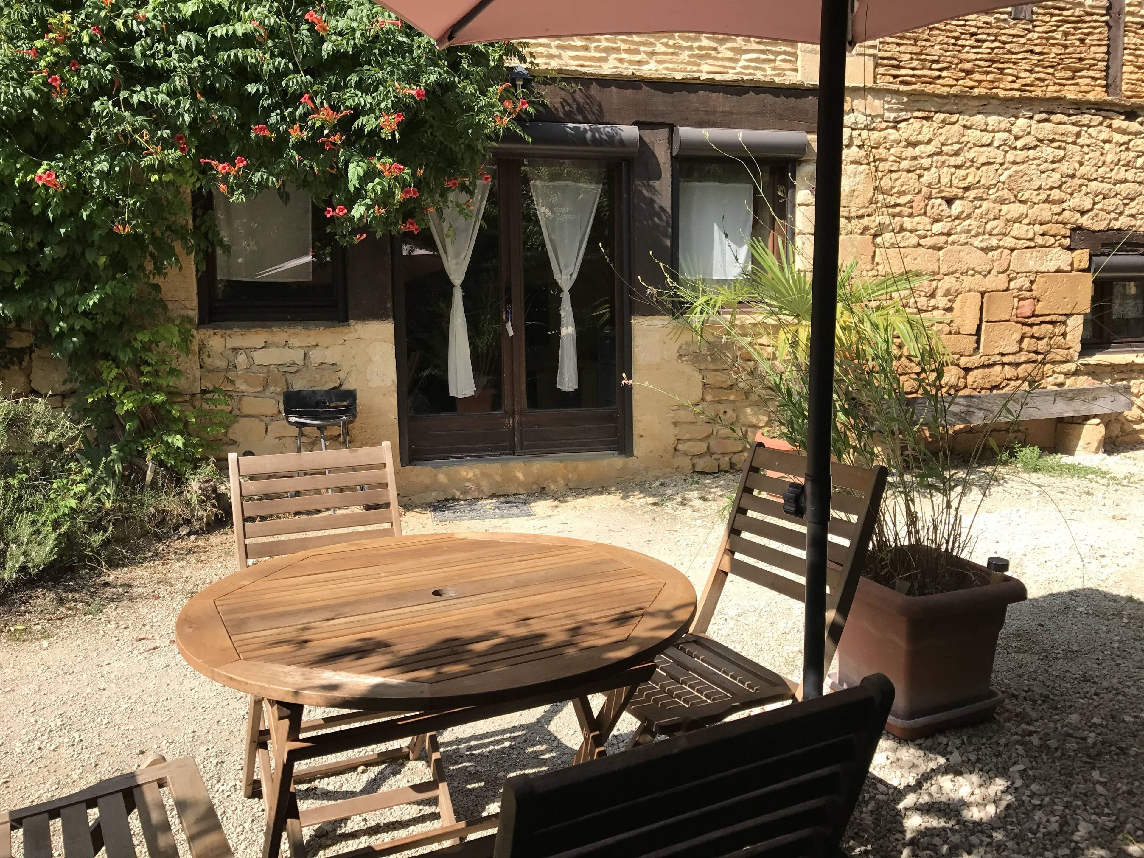 Ein rustikaler Sitzbereich im Freien mit Holztisch und -stühlen, einem Blumentopf und einem Steinhaus mit Fenster und Tür im Hintergrund.