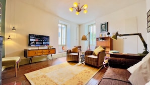 Una sala de estar con una televisión de pantalla plana, un mueble de madera, un sofá a cuadros, una mesita y una lámpara de pie.