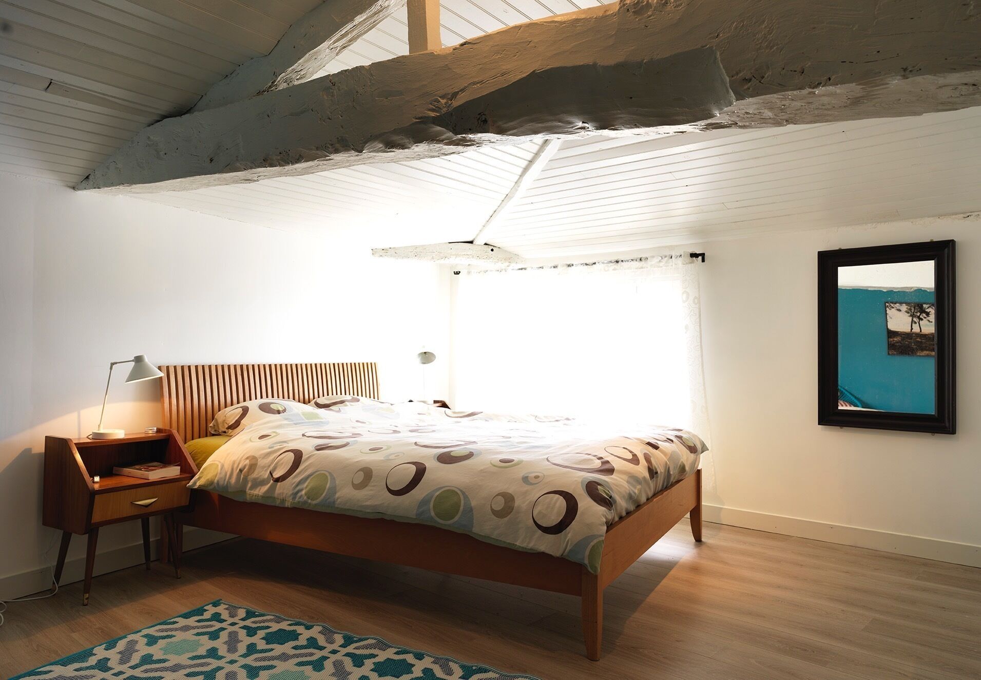 A bedroom with a wooden bed, a nightstand, a lamp, a patterned rug, and a framed picture on the wall.