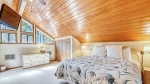 Een slaapkamer met een schuine plafond, een bed met een bloemenbedekking, een nachtkastje met een lamp en een dressoir met lades.