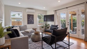 Ein modernes Wohnzimmer mit grauem Sofa, schwarzem Sessel und einem gemusterten Teppich. Zwei Fenster mit Jalousien, ein flachbildfernseher an der Wand und eine Glastür zu einem Balkon.