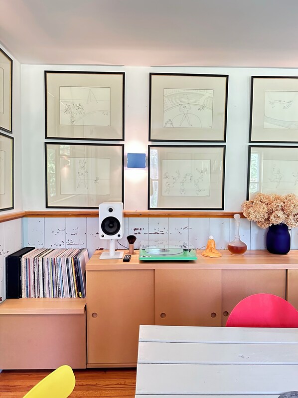 A room with a wooden desk, a speaker, a vinyl record player, and framed sketches on the wall.