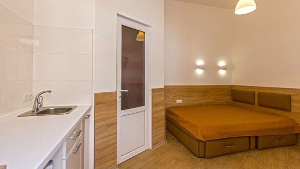 A compact room with a kitchenette, a bed with storage drawers, and a door with a frosted glass panel.