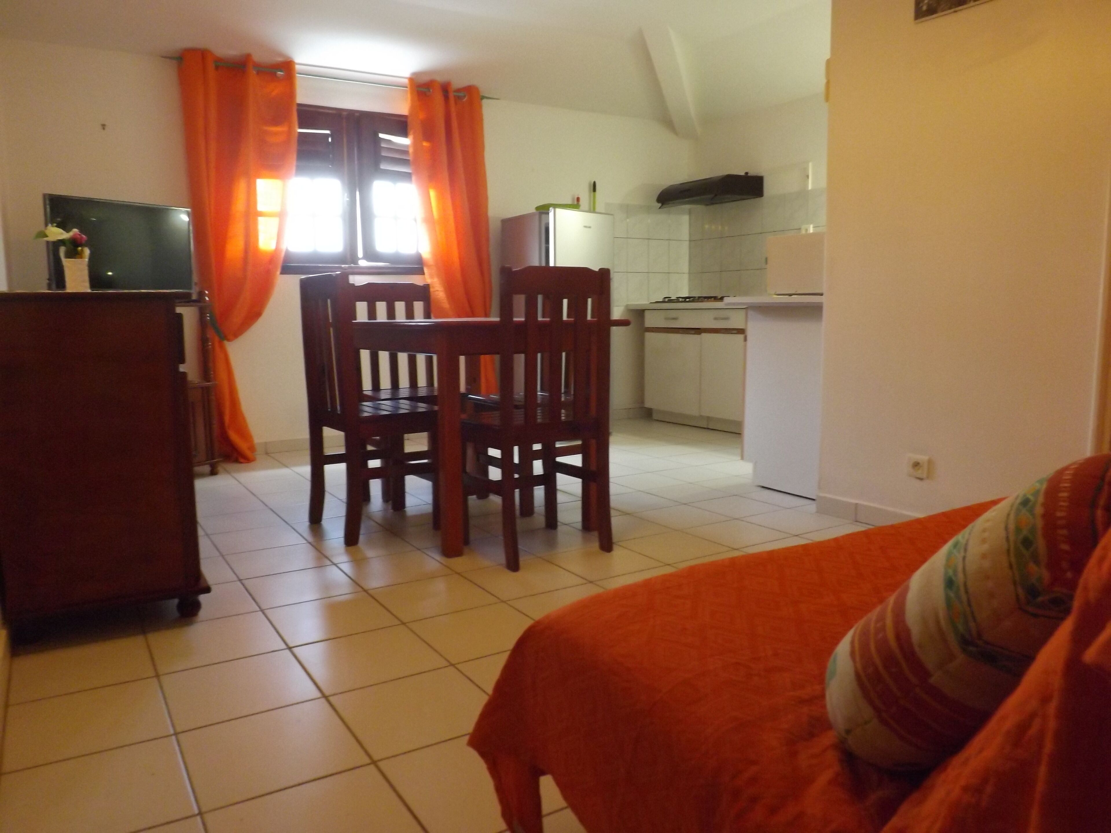 Una stanza con un letto, una sedia in legno, un tavolino, un angolo cottura con pensili e una finestra con tende.
