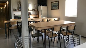 Een eethoek met verschillende tafels en stoelen, een koelkast, een plank met boeken en decoratieve objecten, en een raam met natuurlijk licht.