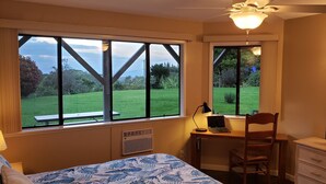 Habitación de hotel con cama, escritorio, silla, televisor y ventanas amplias que dan a un césped y árboles.