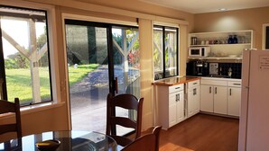 Cocina con alacenas blancas, microondas y refrigerador. Hay una mesa de comedor con sillas y una ventana con vista a un jardín.