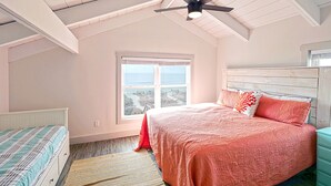Een slaapkamer met een schuine plafond, een bed met een roze sprei, een nachtkastje, een raam met uitzicht en een plafondventilator.