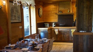 A wooden interior with a dining table set for four, a kitchen area with appliances, and a large window. - Superb chalet in Megève, 5CH, 4 bathrooms, close to center and ski lifts (Megève)