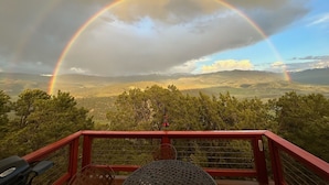 Uitzicht vanaf een balkon met een rode reling, over een landschap met bergen en een regenboog.