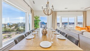 A dining area with a long wooden table set for six, large windows offering a view of buildings and greenery, and a chandelier above.