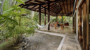 Un espacio al aire libre cubierto con una mesa y sillas, rodeado de plantas tropicales y una hamaca.