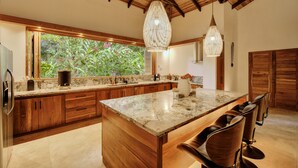 Una cocina amplia con armarios de madera, una isla grande con taburetes de bar y vistas a exuberante vegetación a través de un ventanal.