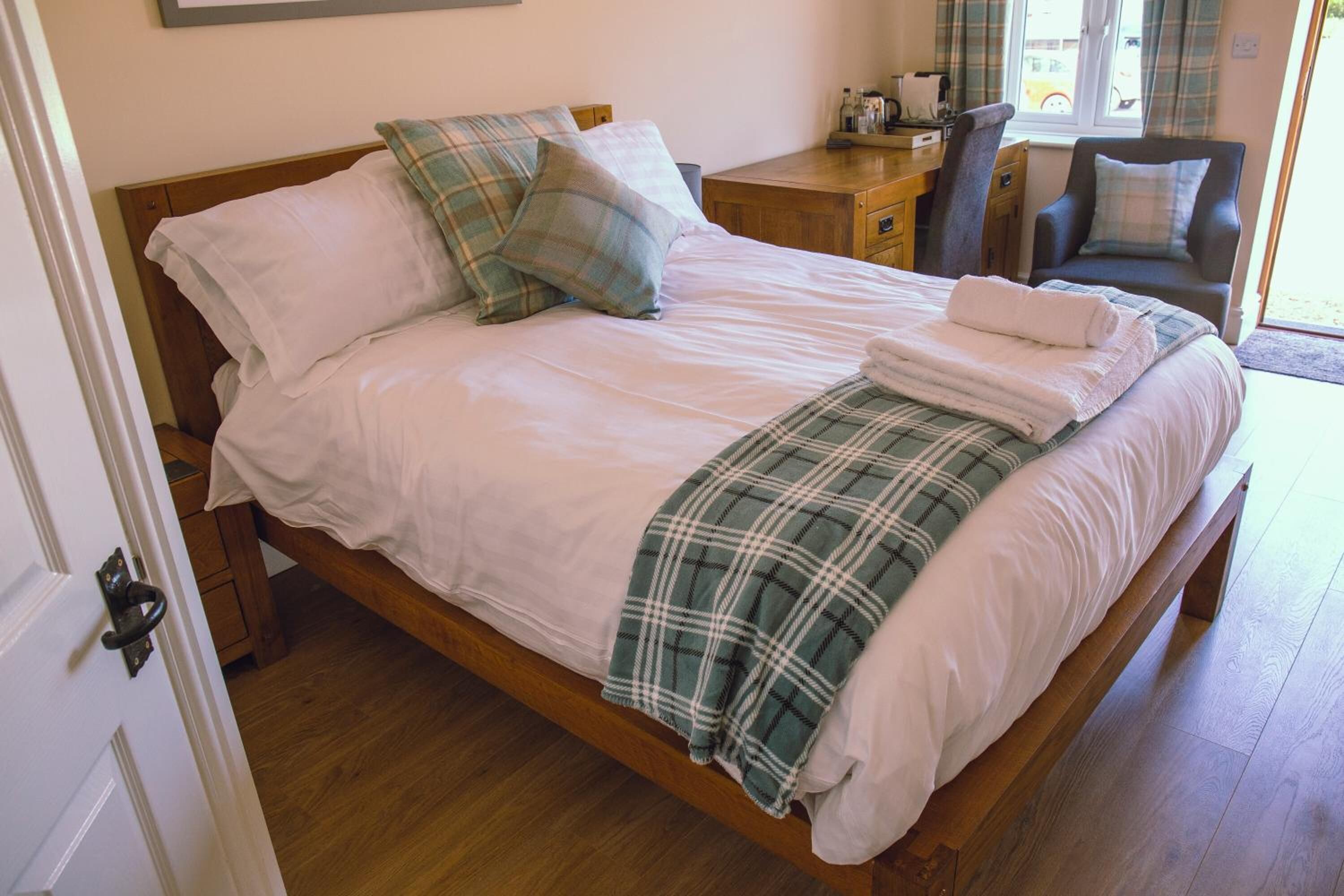 Un lit bien fait, avec une couverture à carreaux et des oreillers, une table de chevet en bois avec une lampe, et une chaise près de la fenêtre.