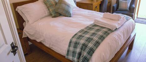 Una cama bien hecha con una manta y almohadas de cuadros, una mesita de noche de madera con una lámpara, y una silla junto a la ventana.