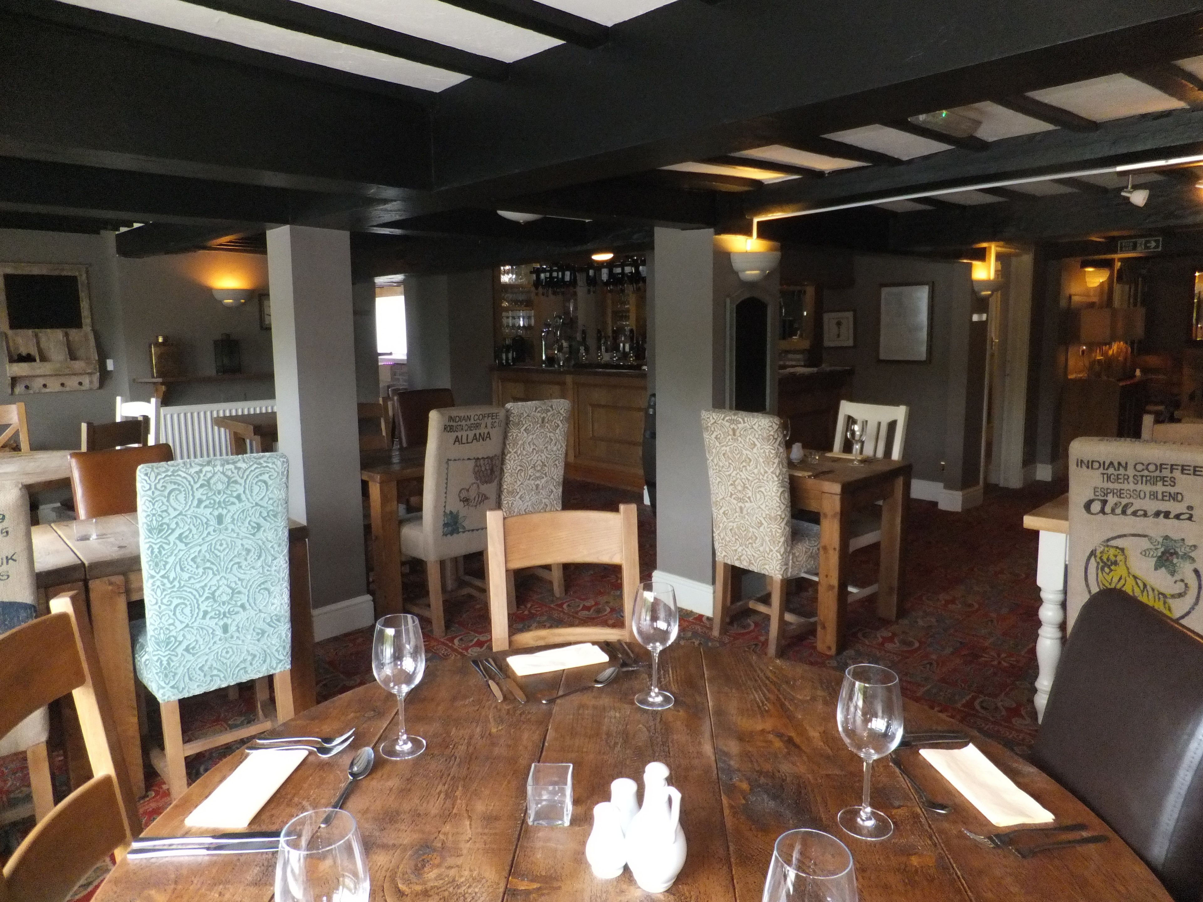A dining area with wooden tables and chairs, a bar in the background, and a patterned tablecloth on one chair.