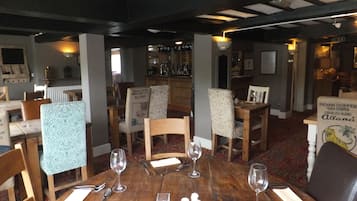 Un área de comedor con mesas y sillas de madera, una barra al fondo y un mantel estampado en una silla.