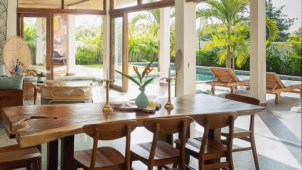 A spacious indoor area with a large wooden dining table, multiple chairs, and a view of a swimming pool and greenery outside.