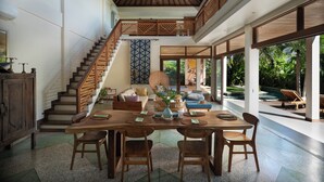 A spacious living area with a wooden staircase, a dining table with chairs, and a view of a pool and greenery outside.