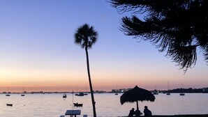 Escena de playa con una palmera, una sombrilla de paja y una mesa con bancos.