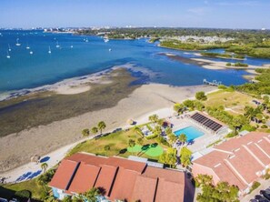 Vue aérienne d’une zone côtière avec une plage, une piscine et un port de plaisance où sont amarrés des bateaux.