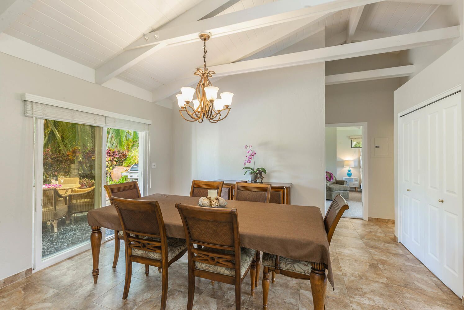 A dining area with a large table, chairs, and a chandelier. There is a sliding glass door leading to an outdoor area with plants.