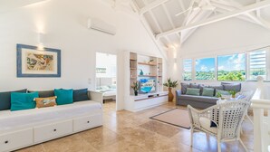 A spacious living area with a white sofa, a wicker chair, and a TV on a white entertainment unit. - Céladon (Le François)