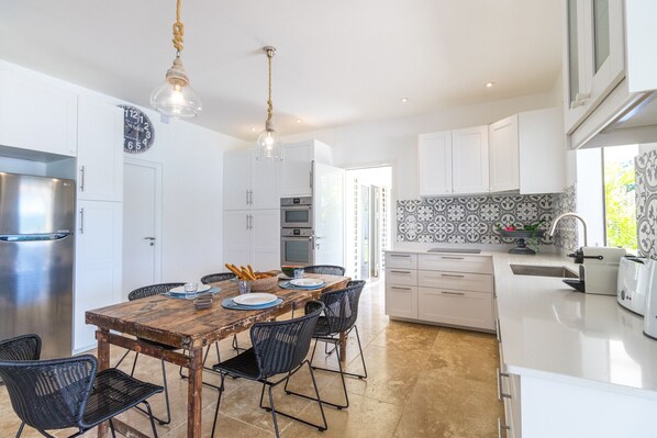 A modern kitchen with white cabinets, a wooden dining table, black wicker chairs, and hanging pendant lights.