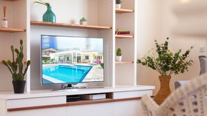 A living room with a flat-screen TV displaying a poolside view, wooden shelves with plants, and a white sofa. - Céladon (Le François)