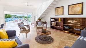 A spacious living area with a view of a pool and outdoor seating, a staircase, and a bookshelf with decorative items. - Céladon (Le François)