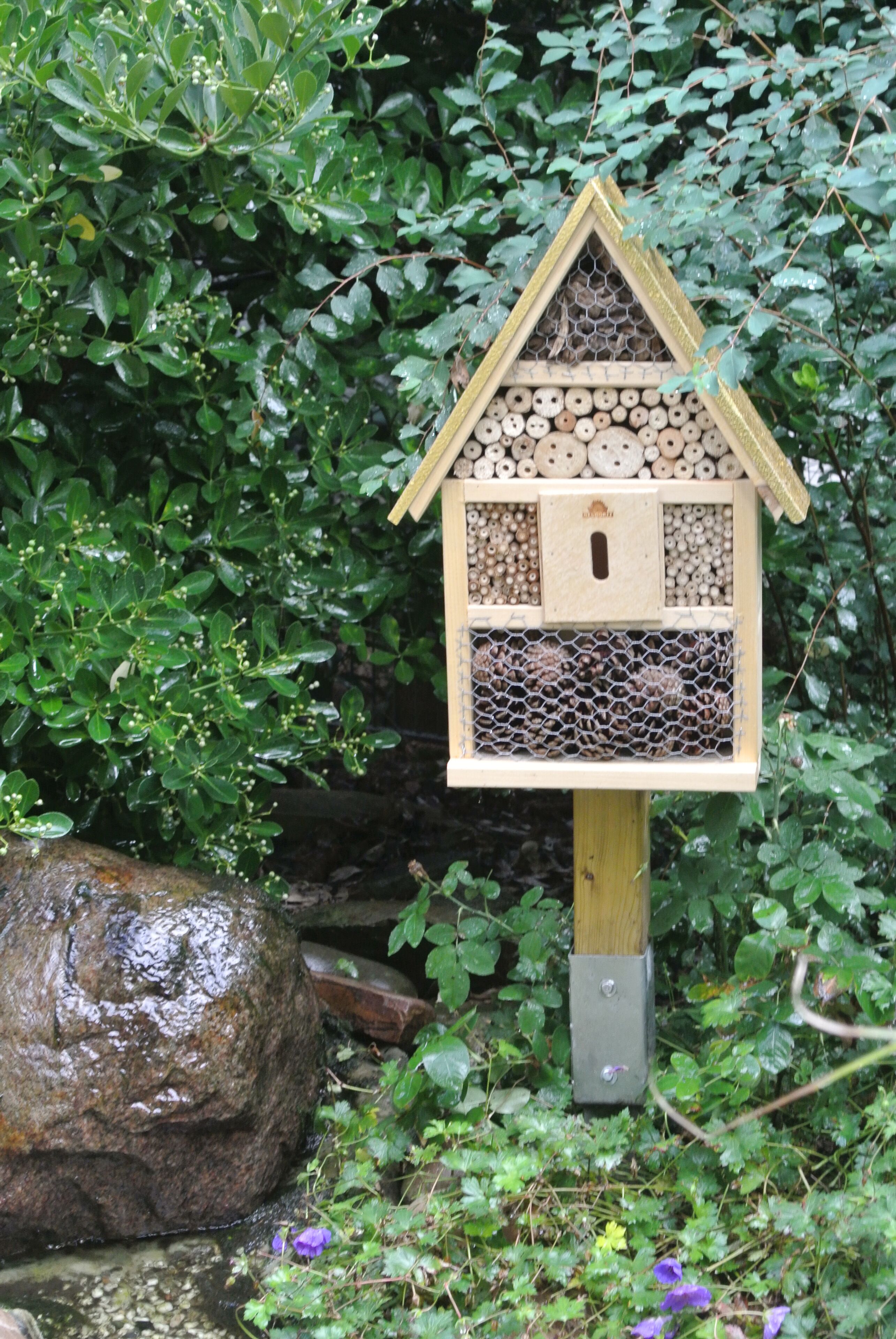 Ein Vogelhaus aus Holz mit Nistfächern vor sattem Grün und einem großen Felsen.