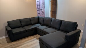 A modern living room with a sectional sofa, wooden flooring, and a door leading to another room. - Blue Bear Hideout the ultimate set up for large groups and get togethers! (Kalkaska)