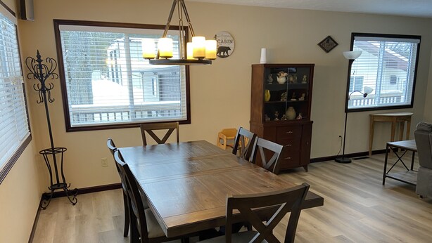 A dining room with a wooden table and chairs, a chandelier, a cabinet with decorative items, and large windows with blinds. - Blue Bear Hideout the ultimate set up for large groups and get togethers! (Kalkaska)