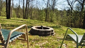 A circular stone fire pit surrounded by grass, with two metal benches facing it. - Blue Bear Hideout the ultimate set up for large groups and get togethers! (Kalkaska)
