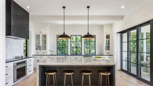 Une cuisine moderne dotée d’un grand îlot central, de tabourets de bar, de luminaires suspendus et de grandes fenêtres offrant une vue sur la verdure.