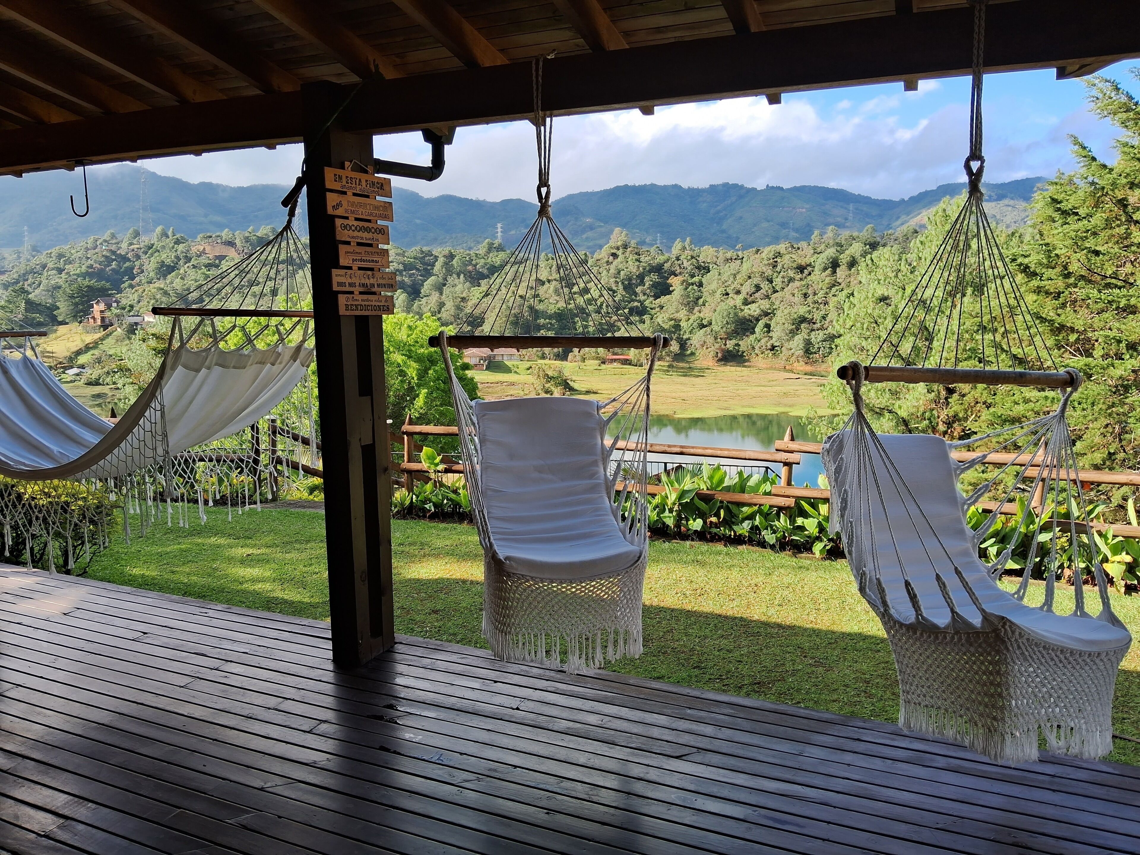 Terraza de madera con tres hamacas blancas y un letrero, con vistas a un paisaje verde exuberante que incluye un lago y montañas a lo lejos.
