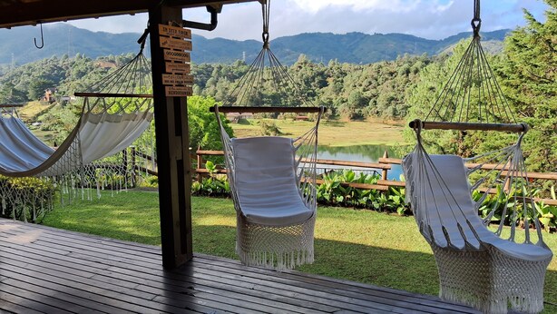 Terraza de madera con tres hamacas blancas y un letrero, con vistas a un paisaje verde exuberante que incluye un lago y montañas a lo lejos.