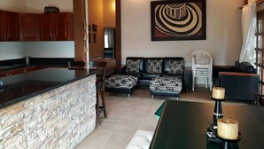 Una sala de estar moderna con una pared de acento de piedra, un sofá negro con cojines estampados, una mesa de comedor de madera y un cuadro abstracto grande en la pared.